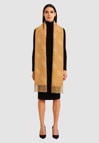 Sciarpa color cammello con motivo geometrico e logo, realizzata in materiale morbido con estremità sfrangiate, indossata sopra un vestito nero con collo alto.