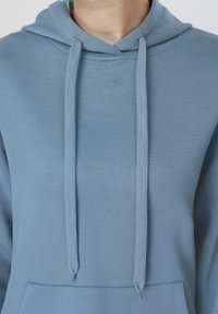 Άτομο που φοράει ανοιχτό μπλε φούτερ με μπροστινή τσέπη και μακριά κορδόνια που κρέμονται στο στήθος, μπροστά σε ουδέτερο φόντο.