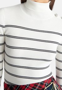 Ribbede striktrøje med høj hals i hvid med sorte vandrette striber, med dekorativ hardware på skulderen.
