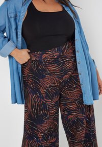Schwarzes Tanktop unter einem hellblauen Jeanshemd, kombiniert mit weiten Hosen in einem dunkelblauen und braunen abstrakten Muster.