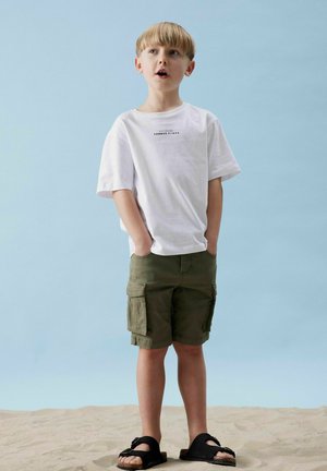 Weißes Baumwoll-T-Shirt mit kurzen Ärmeln und Text, kombiniert mit olivgrünen Cargo-Shorts mit Seitentaschen, schwarzen Sandalen, sandigem Boden.
