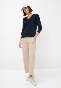 Marineblauer Strickpullover mit V-Ausschnitt, kombiniert mit cremefarbenen weitgeschnittenen Hosen und beigen Sneakern. Das Model trägt eine helle Mütze und eine Halskette.