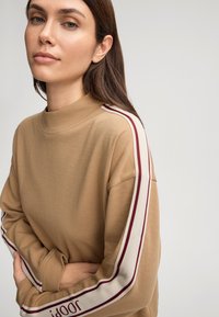Felpa marrone con collo alto; presenta strisce bordeaux e bianche lungo le maniche e dettagli testuali al polsino. Tessuto morbido e textured.