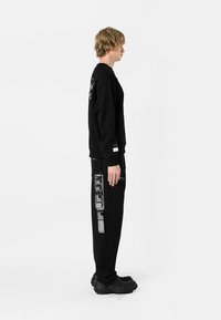 Sudadera negra y pantalones de chándal con texto y gráficos. Los pantalones de chándal presentan un patrón distintivo en el costado. El calzado son zuecos negros texturizados.
