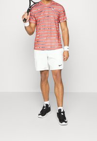 Camisa deportiva a rayas rojas y azules, pantalones cortos blancos con bolsillos, zapatillas negras, sosteniendo una raqueta de tenis, muñequeras blancas, corte atlético.