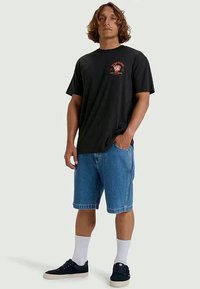 Schwarzes Baumwoll-T-Shirt mit einem roten Grafikdesign auf der Vorderseite, kombiniert mit hellblauen Denim-Shorts und schwarzen Turnschuhen mit weißen Socken.