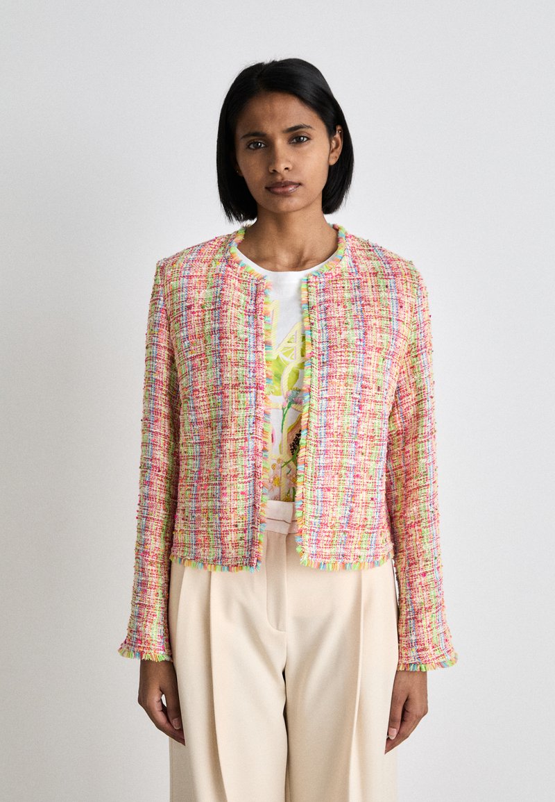 Rosa Tweed-Jacke in cropped Form mit mehrfarbigen Akzenten. Sie hat einen runden Ausschnitt und Fransenverzierungen entlang der Kanten. Getragen über einem bedruckten Oberteil.