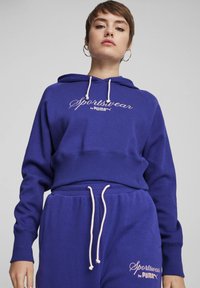 Fialová mikina s kapucňou a skráteným strihom, s bielou výšivkou "Sportswear by PUMA" a doplnenými svetloružovými šnúrkami.