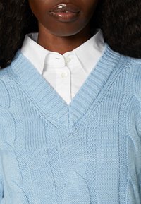 Personne à la peau foncée portant une chemise blanche boutonnée sous un pull en maille torsadée bleu clair à col en V.