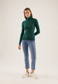 Dark green ribbed turtleneck sweater with long sleeves paired with light blue, slightly cropped jeans and white sneakers.