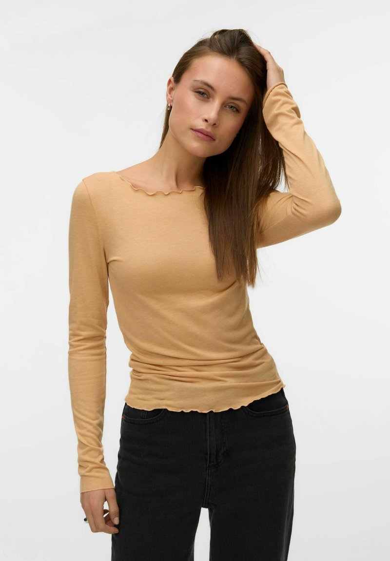 Kvinde med langt brunt hår, iført en tætsiddende beige langærmet top med bølgede kanter og sorte højtaljede jeans, poserende med den ene hånd på hovedet.