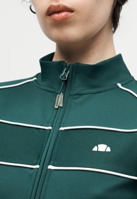 Κοντινό πλάνο ενός ατόμου που φοράει πράσινο μπουφάν με φερμουάρ, λευκές ραφές και μικρό λευκό ημικυκλικό λογότυπο στο στήθος.