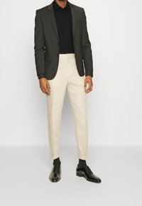 Blazer verde oscuro sobre una camisa negra, combinado con pantalones tapered beige claros y zapatos de vestir negros brillantes. Textura suave en toda la prenda.