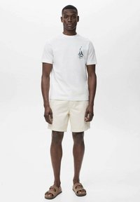 White cotton t-shirt features a small blue sailing boat graphic. Paired with beige shorts and brown sandals. Clean, casual summer outfit.