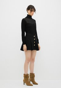 Haut noir à col roulé avec un détail transparent sur la poitrine, associé à une jupe noire évasée ornée de boutons dorés, et des bottines marron à design à fronces.