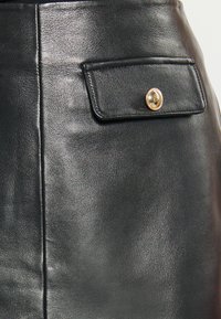 Fustă din piele neagră cu un buzunar frontal dotat cu un buton auriu. Textură netedă cu cusături de-a lungul cusăturii laterale.