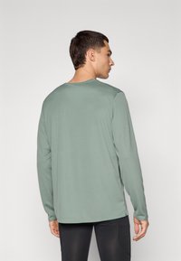 Chemise à manches longues en tissu vert muted, avec un col rond, une coupe ajustée et une texture lisse. Design minimal, sans motifs ni accents.