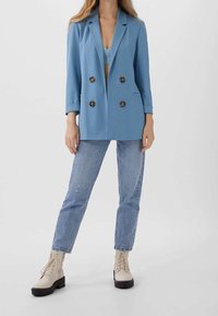 Modrá dvouřadová sako s hnědými knoflíky, spárované se světlými džíny a béžovými šněrovacími botami. Hladká tkanina a strukturovaný design.