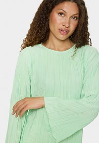 Jasnozielona bluzka z plisami, z długimi, rozszerzającymi się rękawami i okrągłym dekoltem. Materiał wydaje się miękki i teksturowany z subtelnymi liniami pionowymi.