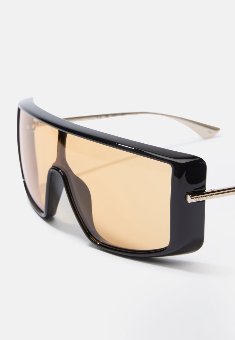 Übergroße schwarze Sonnenbrille mit einem massiven Rahmen. Braun getönte Gläser, goldverzierte Bügel und eine glatte, glänzende Oberfläche.