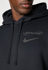 Black hoodie made of soft fabric. Features a gray "SPURS" logo and a Nike swoosh. Includes a drawstring hood and textured detailing.
