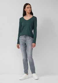 Chemise verte à manches longues, décolleté en V, avec une coupe décontractée, associée à un jean slim gris clair et des baskets blanches. Texture de tissu lisse.