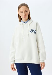 Felpa bianca con cappuccio e testo blu navy "COOL MIND" sul petto, caratterizzata da una vestibilità comoda e polsini a costine. Indossata sopra una camicia con colletto.