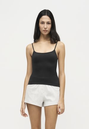 Femme aux longs cheveux foncés portant un débardeur noir et un short gris clair, debout devant un fond blanc uni.