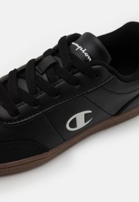 Svart lær sneaker med gummi såle, med svarte lisser og hvit Champion-logo på siden. Glatt tekstur og avslappet design.
