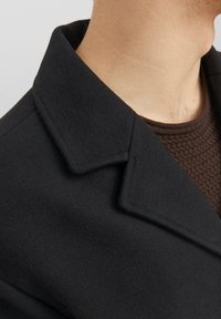 Svart ullkappa med slagkrage och mjuk textur, lager-på-lager över en texturerad brun stickad tröja. Närbild på halsringningen.