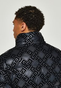 Black puffer jacket with a high collar, featuring a subtle geometric pattern. Glossy finish with a quilted texture.