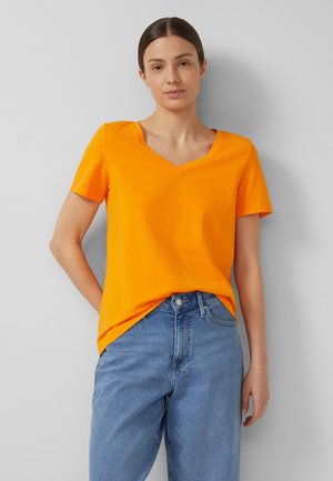 Vrouw met bruin haar in een oranje T-shirt met V-hals en blauwe spijkerbroek, staand tegen een lichtgrijze muur, met één hand rustend op haar heup.
