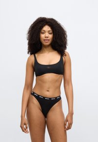 Juoda maudymosi liemenėlė su plačiomis petnešėlėmis ir logotipo detalė centruje, derinama su juodais thong tipo apatiniu, kuriuose yra firminė juostelė juosmens srityje.