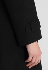 Main reposant à côté de la manche d'un manteau noir avec un détail de sangle en tissu près du poignet.