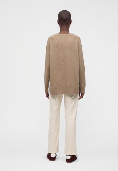 Personne aux cheveux courts, vue de dos, portant un pull beige ample, un pantalon crème et des baskets foncées avec des semelles blanches.