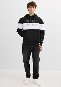 Sudadera con capucha en blanco y negro con gráfico de "EASY DRIP", bolsillo tipo canguro y puños de canalé. Combinada con jeans oscuros y zapatillas blancas.