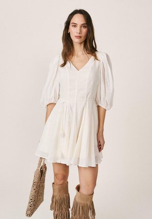 Mujer con vestido blanco de cuello en V y mangas abullonadas, sosteniendo un bolso troquelado con motivo floral en tono marrón claro y botas altas hasta la rodilla con flecos del mismo color.