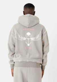 Grijze hoodie met een groot wit grafisch ontwerp met vleugels en een cirkelvormig element op de achterkant, geaccentueerd met roze tekst. Zachte stof, royale pasvorm.