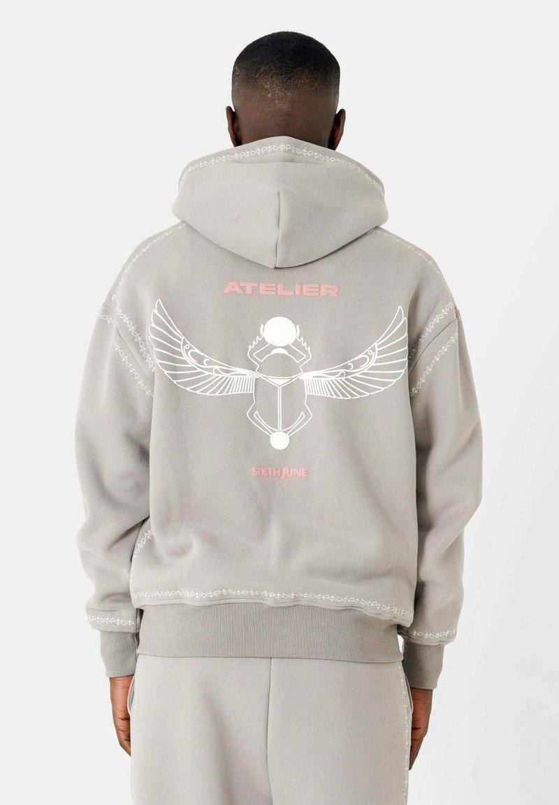 Grijze hoodie met een groot wit grafisch ontwerp met vleugels en een cirkelvormig element op de achterkant, geaccentueerd met roze tekst. Zachte stof, royale pasvorm.