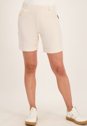 Beige knielange korte broek met omslagzoom, zakken en een gladde tailleband, gedragen met witte sneakers door een persoon die voor een egale achtergrond staat.