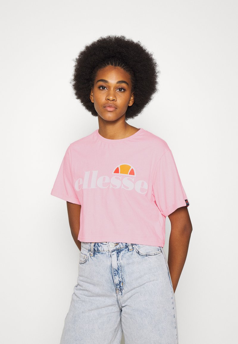 Light pink cropped t-shirt made of cotton, featuring a large gray "ellesse" logo and a colorful half-sun graphic above it. Worn with high-waisted denim shorts.