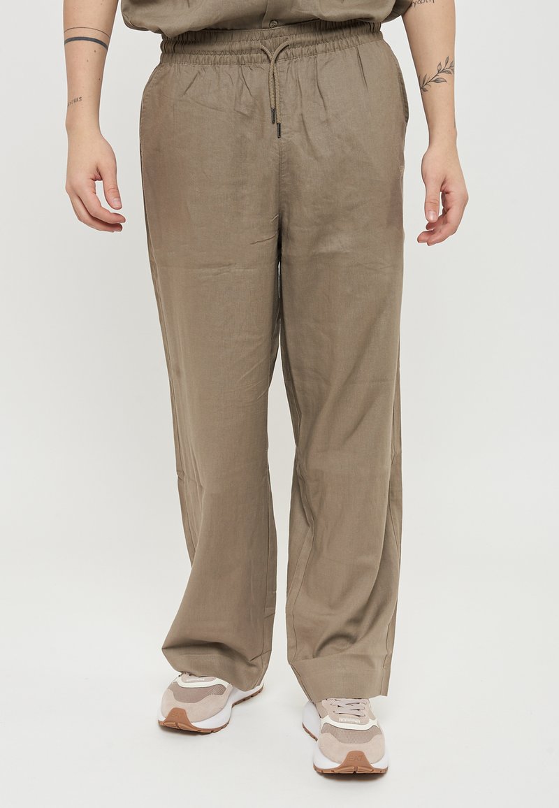 Pantaloni in lino beige leggeri con una vestibilità rilassata, cintura elastica, dettaglio con cordino e minima texture. Indossati con sneakers bianche e beige.
