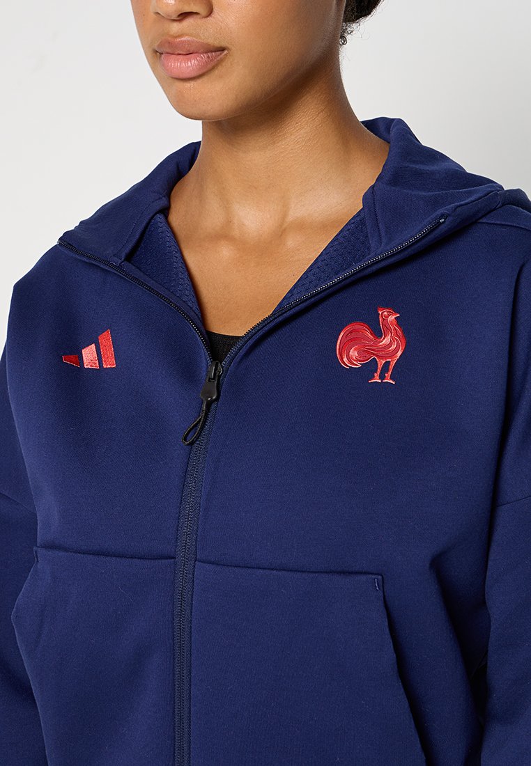 Marineblaue Zip-Hoodie aus weichem Stoff; mit roten Akzenten, einschließlich eines Hahnlogos und drei Streifen auf der linken Schulter.