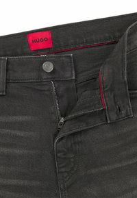 Jeans in denim nero con etichetta rossa, chiusura con zip e bottone argentato. Presentano una texture sottile e dettagli di cucitura sulla vita e sulle tasche.