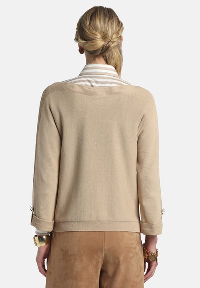 Pull en maille beige à manches trois-quarts, col côtelé et poignets retroussés, superposé à une chemise rayée. Pantalon en daim marron.