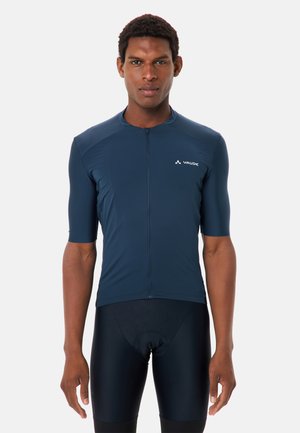 Mann trägt ein marineblaues, kurzärmeliges Radtrikot und schwarze gepolsterte Radhosen, steht vor einem schlichten Hintergrund.