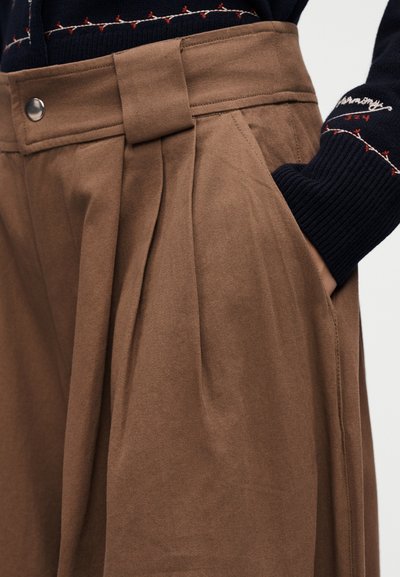 Καφέ παντελόνι ψηλόμεσο και πιετέ με πλαϊνή τσέπη, φορεμένο με μαύρο πουλόβερ με κόκκινη και λευκή κεντήματα στο μανίκι.