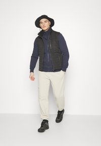 Černá prošívaná vesta přes námořnickou fleecovou mikinu na zip, spárovaná s krémovými teplákovými kalhotami a černými turistickými botami. Klobouk je široký a černý.