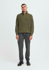 Oliwkowy, prążkowany sweter z rozpinanym zamkiem na ćwierć i wysokim kołnierzem, w połączeniu z szarymi, luźnymi spodniami i czarnymi butami.