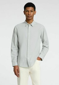 Lichtgrijze langemouwen shirt met een knoopsluiting aan de voorkant, puntige kraag en een gladde textuur. Model draagt crèmekleurige broek.